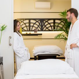 foto de una pareja joven en albornoz, a punto de recibir un masaje en una cabina vip doble. Estan apoyados en la pueda en cada lado de la cabina y se están mirando con expresión de felicidad