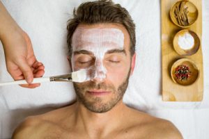 Foto a color de un hombre recibiendo un tratamiento facial. tiene los ojos cerrados y se le esta aplicando la mascarilla con un pincel blanco