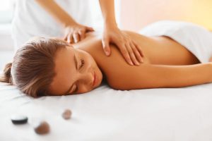 Foto a color de una mujer joven y rubia recibiendo un masaje relajante por la espalda. Esta tumbada boca a bajo, y una terapeuta tiene sus manos apoyadas en su espalda