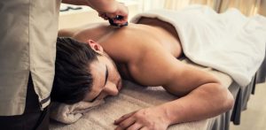 foto a color de un hombre moreno, tumbado en una camilla de masaje. una terapeuta le esta dando un masaje de piedras calientes en la espalda