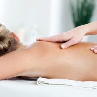 Mujer rubia recibiendo un masaje de espalda tiene el pelo recogido y esta mirando del lado opuesto a la camara. se ven las manos de una masajista deslizando sobre su espalda descubierta