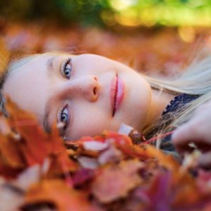 Rostro de una mujer tumba en el suelo sobre hojas rojas de otoño. es Runia tiene los ojos azul y tiene una media sonriza
