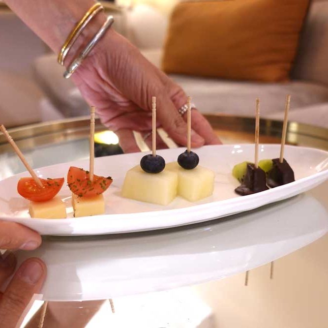 manos de una mujer apoyando una bandeja blanca con una degustación de seis unidades. tomate cherri con queso, melon con arándanos, y kiwi con chocolate. Detrás se ve un sofa gris con dos cojines de color terracota