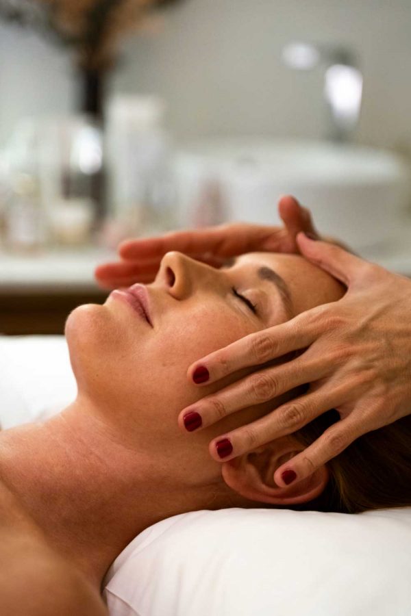 Foto de una mujer rubia, recibiendo un masaje facial relajante. Las manos de la especialista están apoyadas sobre el rostro del modelo. Tiene las uñas pintadas de rojo. En el fondo de la foto se ven unos productos de cosmética desenfocado y un lavabo blanco