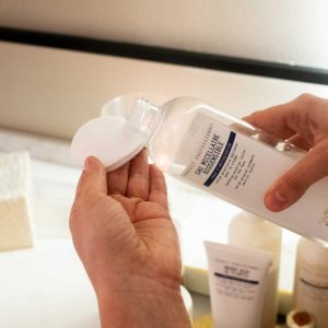 Manos de la terapeuta aplicando eau micellaire sobre un algodón. El bote es transparente y se ven productos de cosmética disfuminados