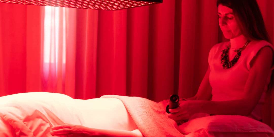 Luz-NIR-Wellness-Boutique placación de luz NiR y Luz Roja en tratamiento . Se ve una placa de luz NIR de color Rojo ecima de una camilla mientras una cliente se esta haciendo un tratamiento facial. tiene el abdomen descubrierto , y Maria casado le esta aplicando 2 laseres Lyma en el rostro. en la parte traser se ve una cortina beige. la sala esta iluminada de color rojo, debido a la luz NIR