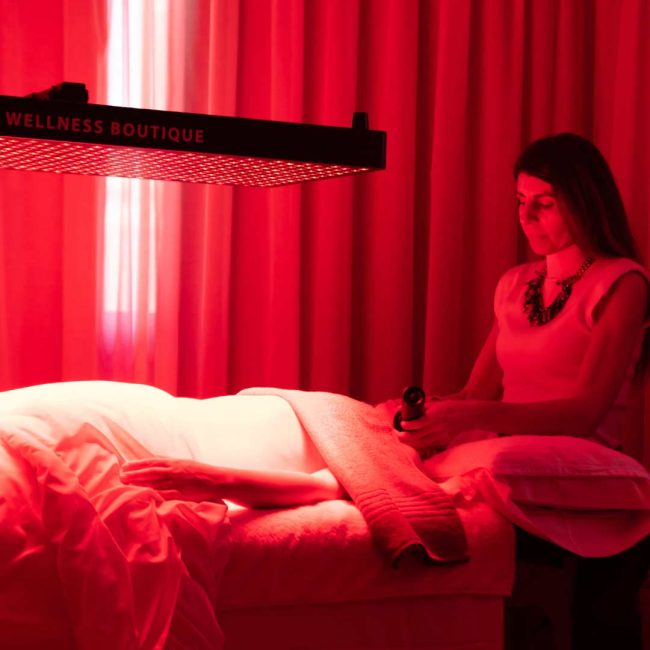 Luz-NIR-Wellness-Boutique placación de luz NiR y Luz Roja en tratamiento . Se ve una placa de luz NIR de color Rojo ecima de una camilla mientras una cliente se esta haciendo un tratamiento facial. tiene el abdomen descubrierto , y Maria casado le esta aplicando 2 laseres Lyma en el rostro. en la parte traser se ve una cortina beige. la sala esta iluminada de color rojo, debido a la luz NIR