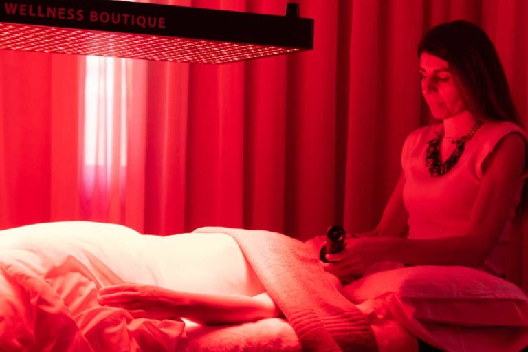 placación de luz NiR y Luz Roja en tratamiento . Se ve una placa de luz NIR de color Rojo ecima de una camilla mientras una cliente se esta haciendo un tratamiento facial. tiene el abdomen descubrierto , y Maria casado le esta aplicando 2 laseres Lyma en el rostro. en la parte traser se ve una cortina beige. la sala esta iluminada de color rojo, debido a la luz NIR